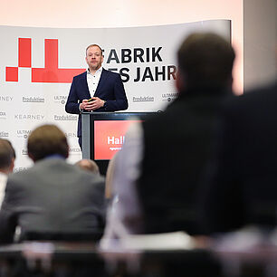 FABRIK DES JAHRES - Tobias Hallmann steht auf der Bühne hinter dem Rednerpult und spricht Richtung Publikum